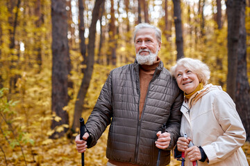 Mature caucasian couple with nordic sticks walking in autumn park, enjoying the weather together. Man and woman with gray hair having rest, wearing casual warm clothes. leisure, healthy lifestyle