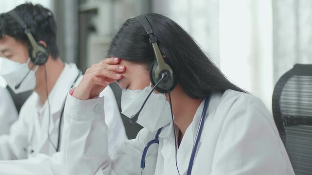 Close Up Of An Asian Woman Doctor Wearing Headsets And Masks Working As Call Center Agent Is Tired Due To Working While Her Colleagues Are Speaking And Typing During A Call At The Office
