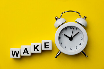 White analog alarm clock isolated on yellow background with word Wake on white plastic cubes. Concept of time management. A clock with the word WAKE. Copy space. Close-up.