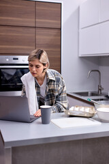 Woman baking at home following recipe on laptop, in light interior kitchenm wearing casual checkered shirt, caucasian beautiful woman is engaged in baking cooking. food ingredients on table.