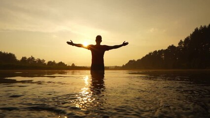 A man stands in the river with his arms open to meet the sun. The man stretched out his arms toward the sun. Calmness, wisdom, and faith with him