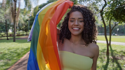 Young curly hair woman covering with lgbt pride flag. Keeping fist up, covering LGBT+ flag