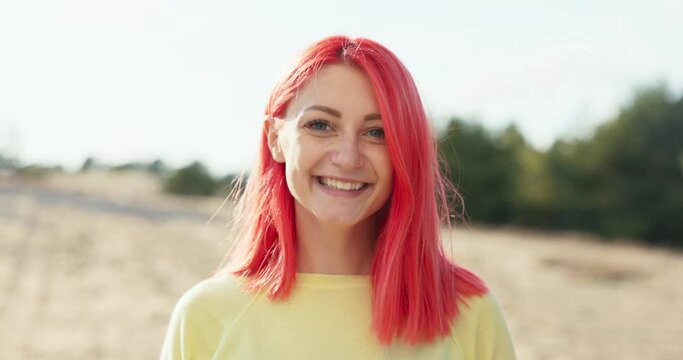 Shot of face smiling beautiful girl with pink hair blowing in wind which she pulls back behind her ear, freckles on face, long thick eyelashes woman is standing on beach among trees, nature outside
