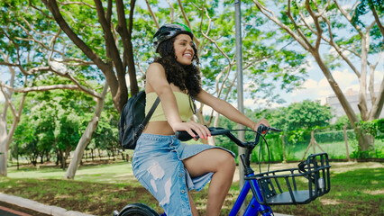 Young Latin woman in protective helmet is riding her bicycle along the bike path in a city park