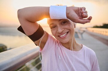 Headshot of a beautiful Hispanic female athlete wiping sweat from her forehead with terry wristband, smiles with cheerful toothy smile resting after morning jog on a city bridge treadmill at sunrise