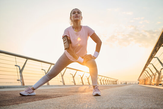 Determined sportswoman with healthy body doing side lunges, stretching her legs muscles while working out outdoor on a city bridge at dawn. Sport, fitness, body care and active lifestyle concept