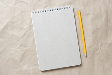 Gray notepad with white coiled spring and pencil on a background of beige crumpled craft paper. With empty space for text and design