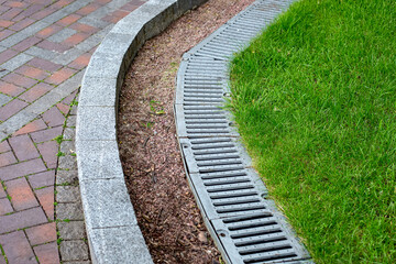 iron drainage grate on the roadside with green grass and stone pebbles at the granite curb along the pedestrian pavement of brick tiles, nobody. - obrazy, fototapety, plakaty