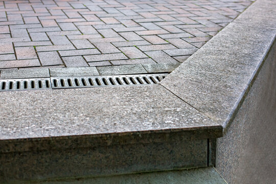 Pavement drainage grate with line iron grill on rectangular stone tile walkway on edge of cliff, close-up of drainage system on dry weather, nobody.