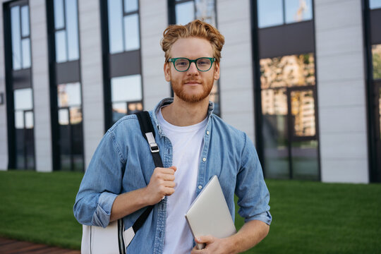 Smiling university student with backpack and laptop computer walking in university campus, education concept. Portrait of confident man freelancer wearing eyeglasses looking at camera on the street 