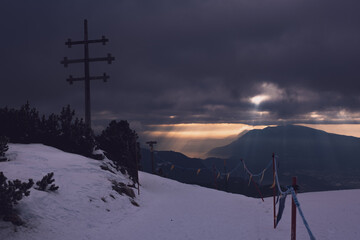 Fototapeta na wymiar Sonnenlicht sticht durch die Wolken in ein Tal vor Bergen mit Schnee und Gipfelkreuz | Ritten, Italien