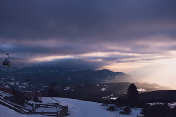 Sonnenlicht fällt durch die Wolken in ein Tal vor Bergen mit Schnee und einer Seilbahn mit Gondel...