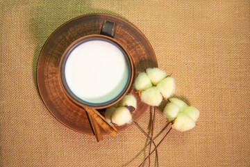 A mug of milk on a saucer with cinnamon and cotton flowers on a table covered with burlap. Top view. Pottery on the table.
