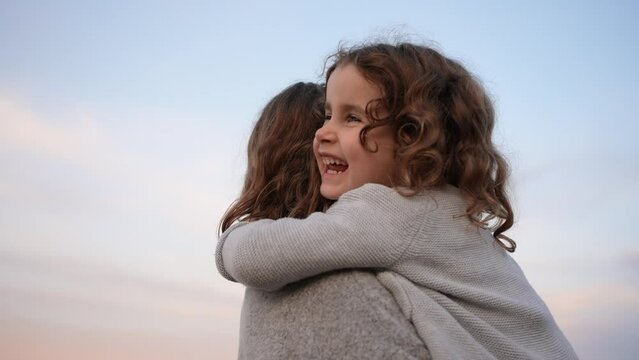 Happy little girl with curly hair gives a hug to her sister and laughs on the beach at sunset. Concept of happy childhood and love between siblings