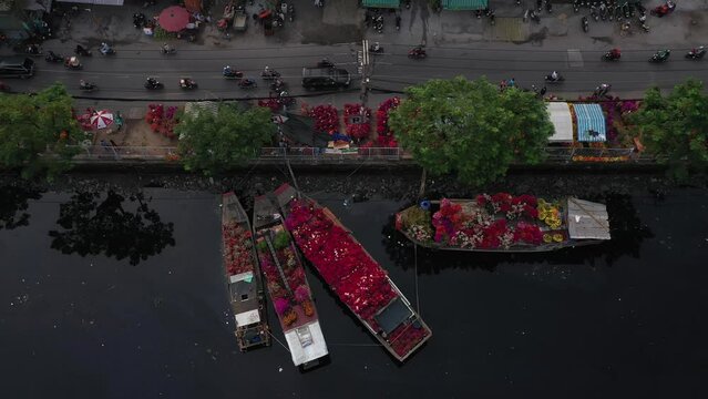 Vietnamese Lunar New Year floating flower market in Ho Chi Minh City is a highlight of the Tet festival from tracking drone view. Farmer Boats with flowers line the busy urban canal in morning light.
