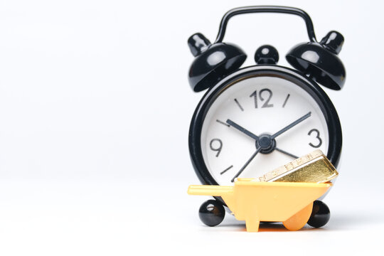 Selective focus with noise effect picture of miniature goldbar own wheelbarrow, alarm clock insight and copyspace white background. Gold investment concept