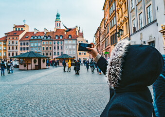 Obraz premium Young Woman in a Fur Hooded Coat Takes a Photo on Phone of Old Town Market Place in Warsaw Poland