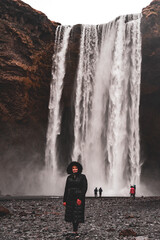 Woman in Black coat with fur hood, yellow hat and orange gloves enjoying Skogafoss Waterfall in...