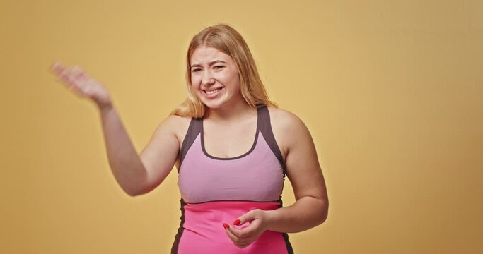 Plus size woman regretting mistake. Chubby blond female in pink sportswear slapping forehead and scratching head after making mistake against yellow background