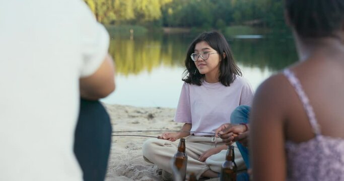 Beautiful woman with glasses sits on the sand near the lake, holds a sausage stick over the campfire, fries it thoroughly on both sides, spends time with friends