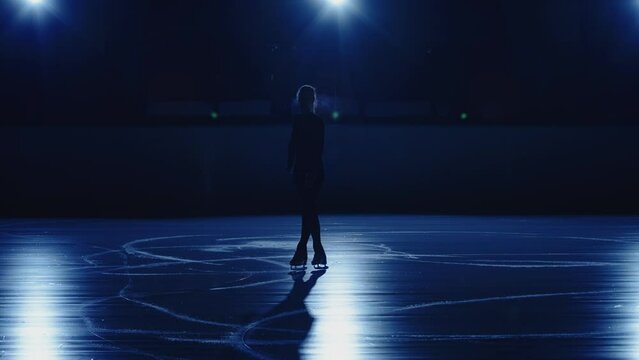 Young female figure skater is performing single skating choreography on ice rink against background of soft blue light. Silhouette of an athlete sliding on ice practicing jumps and spins. Slow motion.