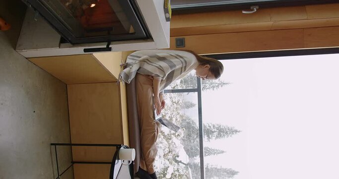 a girl in glasses sits near the window with a laptop on her knees and types then looks out the window the backdrop of a winter forest and next to a burning fireplace with firewood. vertical video