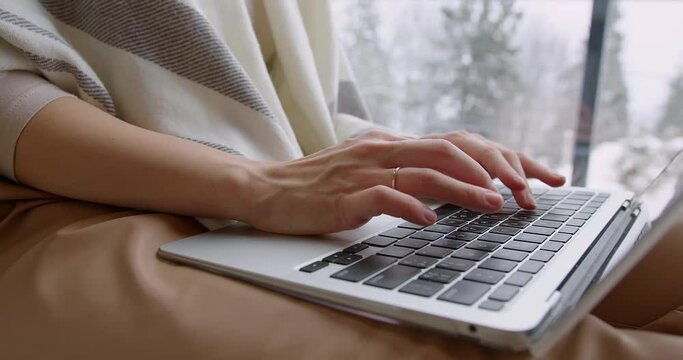 beautiful female hands press the keys of a gray laptop near the window against the backdrop of winter mountains. a girl in a blanket works at a computer sitting on a windowsill. close-up