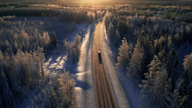 Drone aerial of a car driving on icy road towards sunset in winter. Winter travel and road trip.