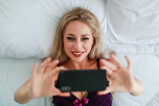 Top view closeup shot of Caucasian blonde hair hot female fashion model in black purple sexy lingerie underwear laying down smiling on bed holding using smartphone taking selfie photo in bedroom