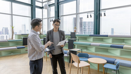 Two young male startup owners have a conversation to exchange ideas in a green co-working space with skyscraper background. Young business workers in casual discuss about their business growth