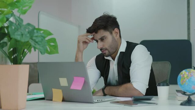 Young Indian man office worker sitting at workplace holding hands massages forehead and head with severe pain. Asian male at work with a headache. Up close face of sufferer is overloaded with sick.