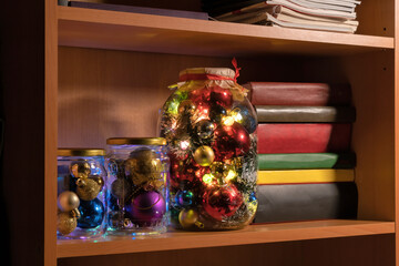 In the bookcase there are glass jars filled with multi-colored Christmas decorations. The lights of the garlands are burning. Background. Celebration.