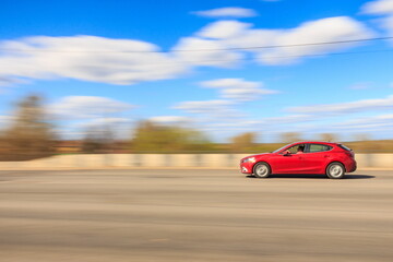 A red car is driving fast on the road on a sunny summer day, the car is in focus, the background is blurred.