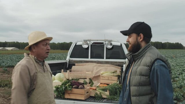 Modern mature Asian farmer selling vegetables to young Caucasian man telling him something then shaking hands