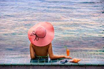 Back view of young woman in pink hat and swimsuit sunbathing at the pool side