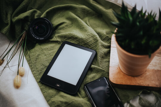 Ebook reader on a green tablecloth. Amidst a flower pot, a cell phone and a camera lens.