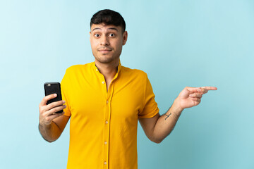 Young Colombian man using mobile phone isolated om blue background surprised and pointing finger to the side