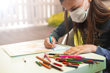 A child in a medical mask draws with colored pencils at a table in a bright room. Place for text