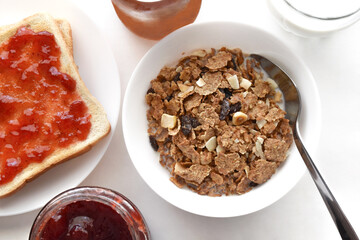 Fruit and fiber cereal and toast with strawberry jam, glass jar of strawberry jam, glass jar of orange jam and glass of milk on the table. Healthy breakfast concept.   Flat lay top view photo.  