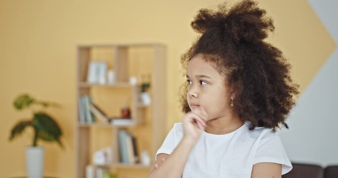 Cute little mixed race girl with dark kinky hair gets confused and uncertain sitting and looking around in children room close view