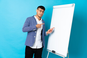 Young Colombian man isolated on blue background giving a presentation on white board and surprised while pointing side