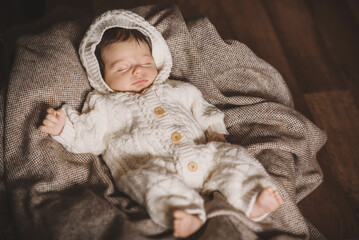 A portrait of little newborn in white cozy knitted overall laying on beige woollen plaid. Sleeping...