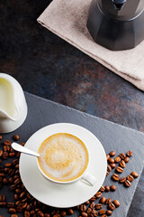 Coffee and coffee beans on a slate board. Cup of coffee on a white saucer and a milk jug on a dark background