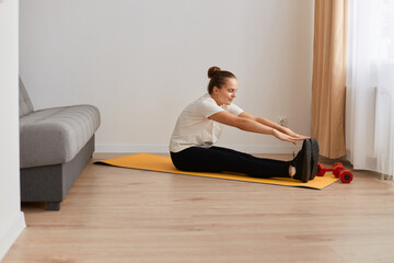 Full length profile portrait of beautiful athletic young woman practicing yoga at home, sitting on mat and stretching her legs, doing sport exercises for her health, fitness and lifestyle.
