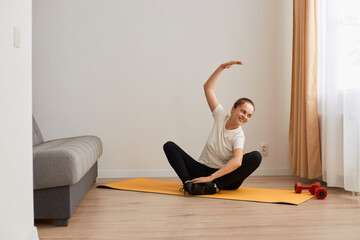 Full length portrait of young healthy beautiful woman in T-shirt and leggings practicing yoga at home sitting in lotus pose on yoga mat and tiling her body to side, stretching.