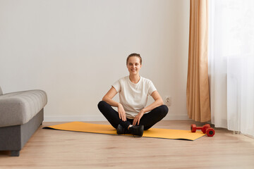 Fototapeta premium Portrait of attractive sporty woman sitting in lotus pose on mat, wearing white t shirt and black leggins, looking at camera with happy smile, meditating and training at home in living room.