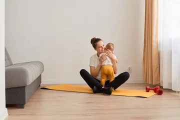 Tuinposter Full length portrait of sporty woman sitting in lotus pose on mat, wearing white t shirt and black leggins, training at home in living room and hugging her baby daughter while having rest. © sementsova321