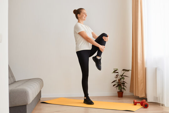 Side View Of Woman Exercising By Lifting The Legs, Knees Up One By One, Standing Straight Back And Hold, Relaxing Yoga Practice At Home, Wearing White T Shirt And Black Leggins.