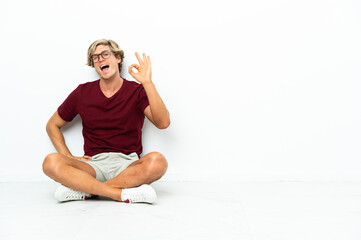 Fototapeta premium Young English man sitting on the floor showing ok sign with fingers
