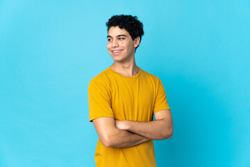 Young Venezuelan man isolated on blue background with arms crossed and happy